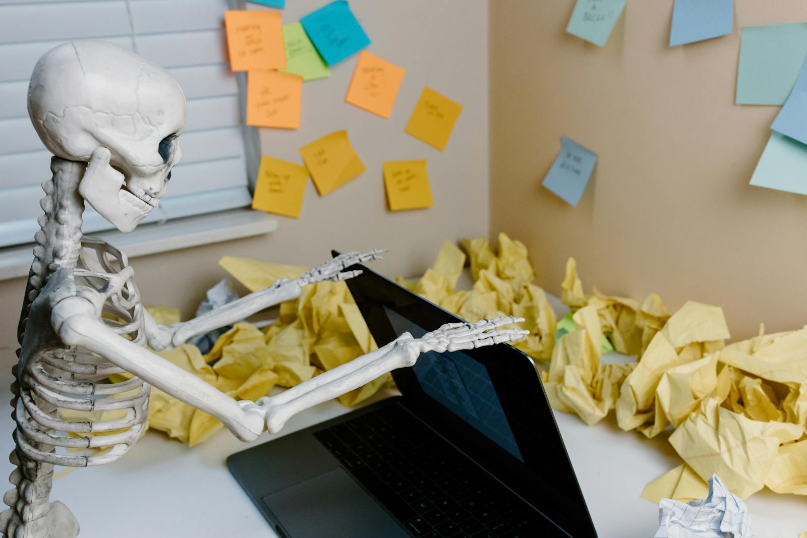 A skeletal figure works at a desk with a laptop and scattered papers, symbolizing burnout.