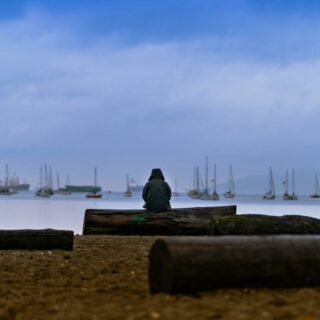 A lone figure sits on a sandy beach in Vancouver, gazing at boats and the calm ocean under a dusky sky.