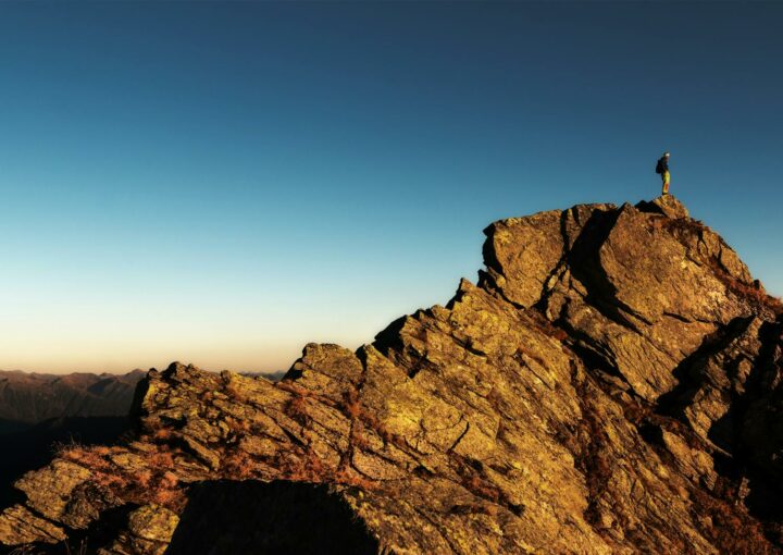 A lone hiker stands triumphantly atop a rocky mountain peak at sunrise, embracing nature and the view.