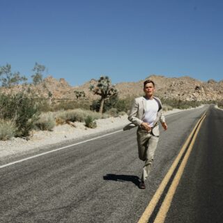 A man in a suit running on an empty desert road surrounded by mountains and dry vegetation under a clear blue sky.