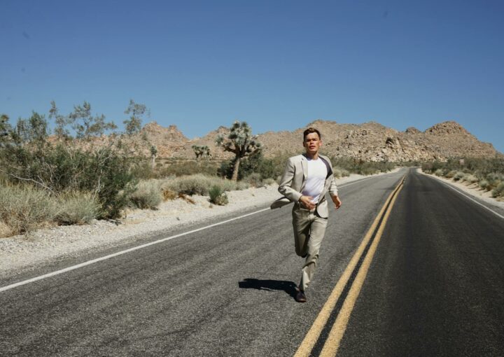 A man in a suit running on an empty desert road surrounded by mountains and dry vegetation under a clear blue sky.