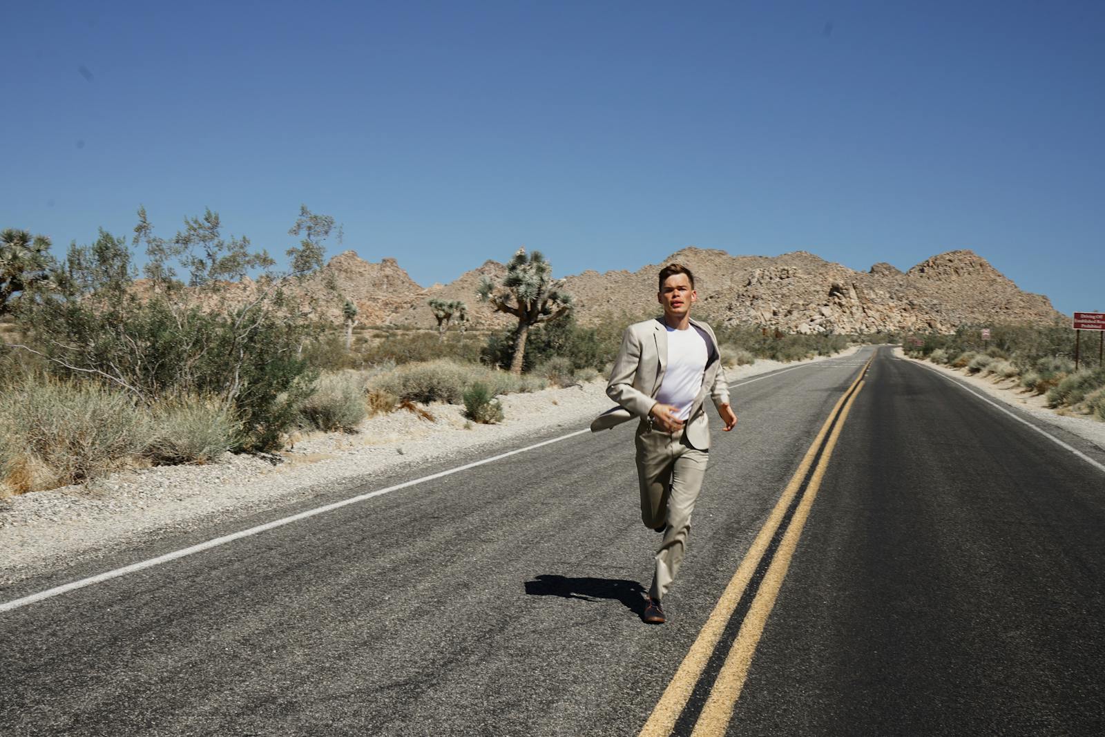 A man in a suit running on an empty desert road surrounded by mountains and dry vegetation under a clear blue sky.