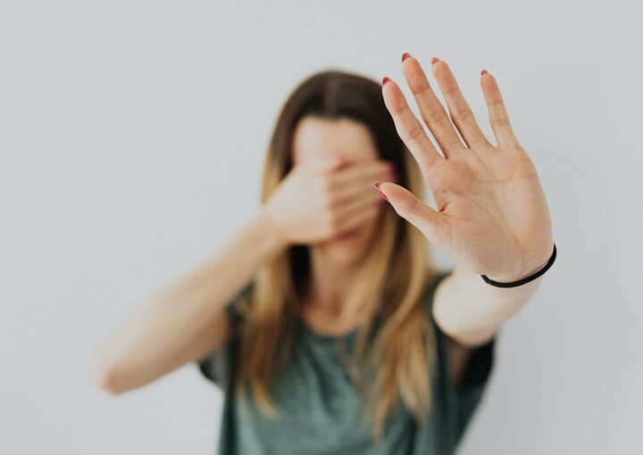 A woman in a gray shirt covers her face with her hand in a stop gesture, evoking a sense of fear and protection.