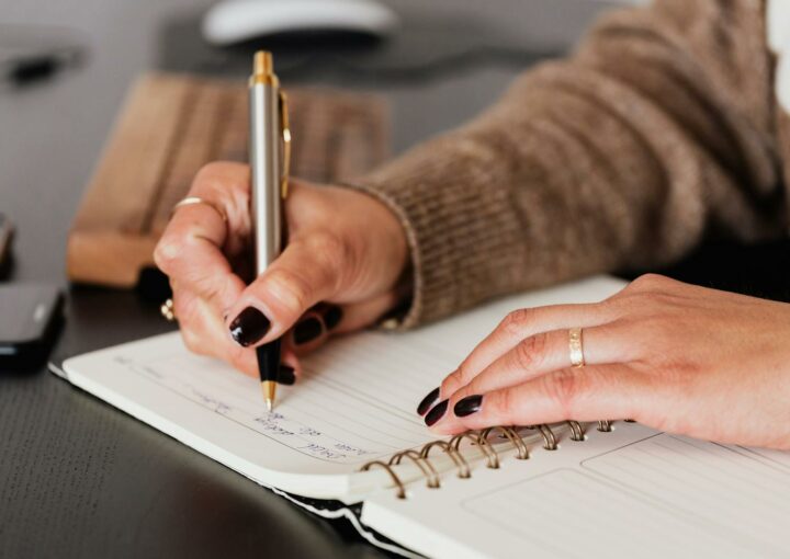 Crop unrecognizable female with stylish manicure sitting at black desk with keyboard and smartphone and taking notes with silver pen in notepad