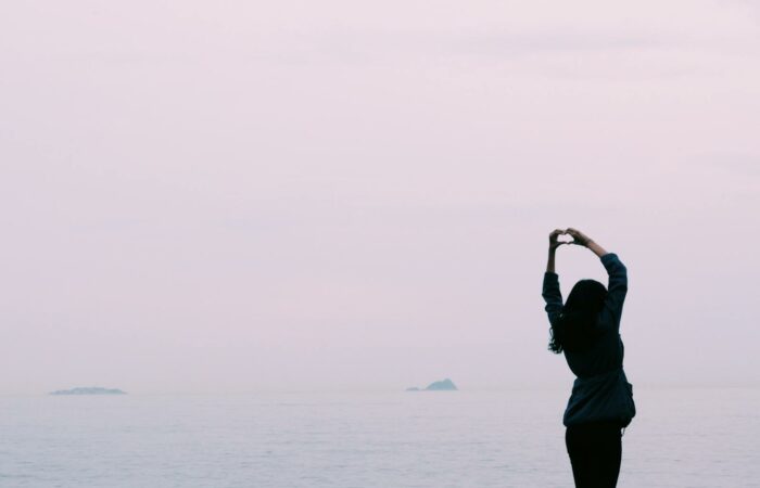 A silhouette of a woman making a heart shape with her hands by the calm sea during sunset.