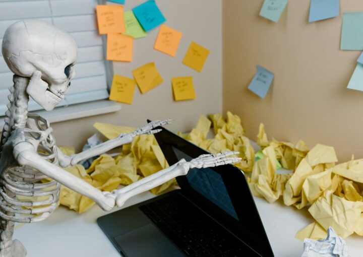 A skeletal figure works at a desk with a laptop and scattered papers, symbolizing burnout.