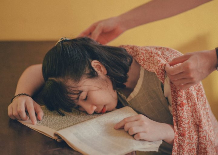 A young woman falls asleep on a book as someone covers her with a blanket.