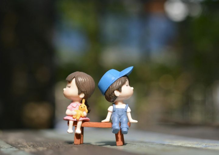 Adorable figurines of a boy and girl sit on a bench, bathed in summer sunlight.