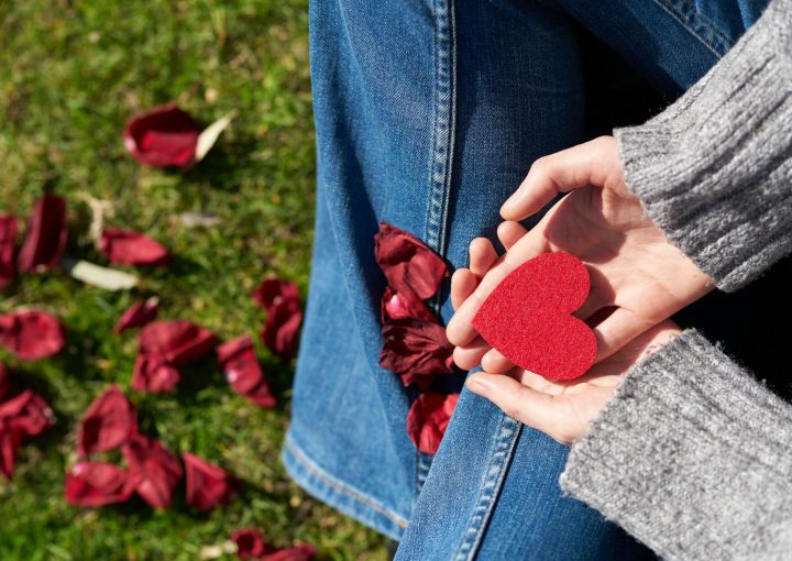 A woman sits on grass holding a red heart-shaped card, surrounded by rose petals, symbolizing love and compassion.