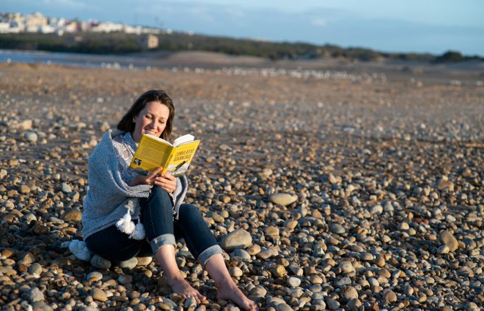 woman in blue denim jacket sitting on rocky shore reading book during daytime