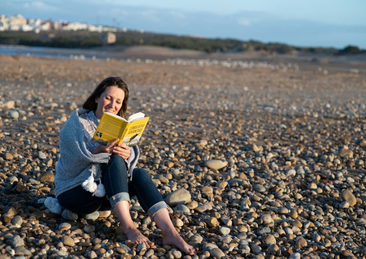 woman in blue denim jacket sitting on rocky shore reading book during daytime