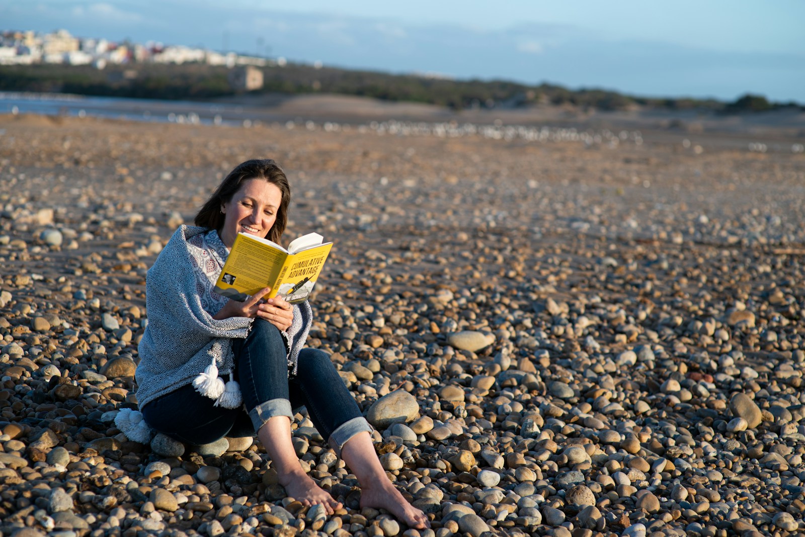 woman in blue denim jacket sitting on rocky shore reading book during daytime