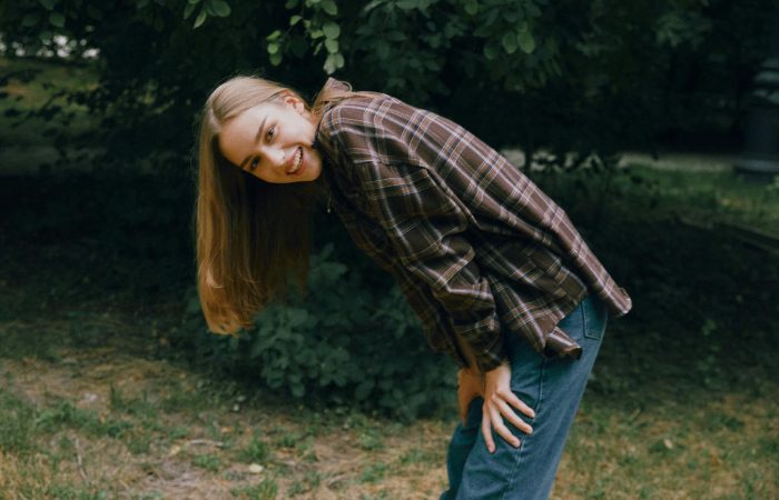 A joyful young woman with long hair bends and smiles outdoors in casual attire.