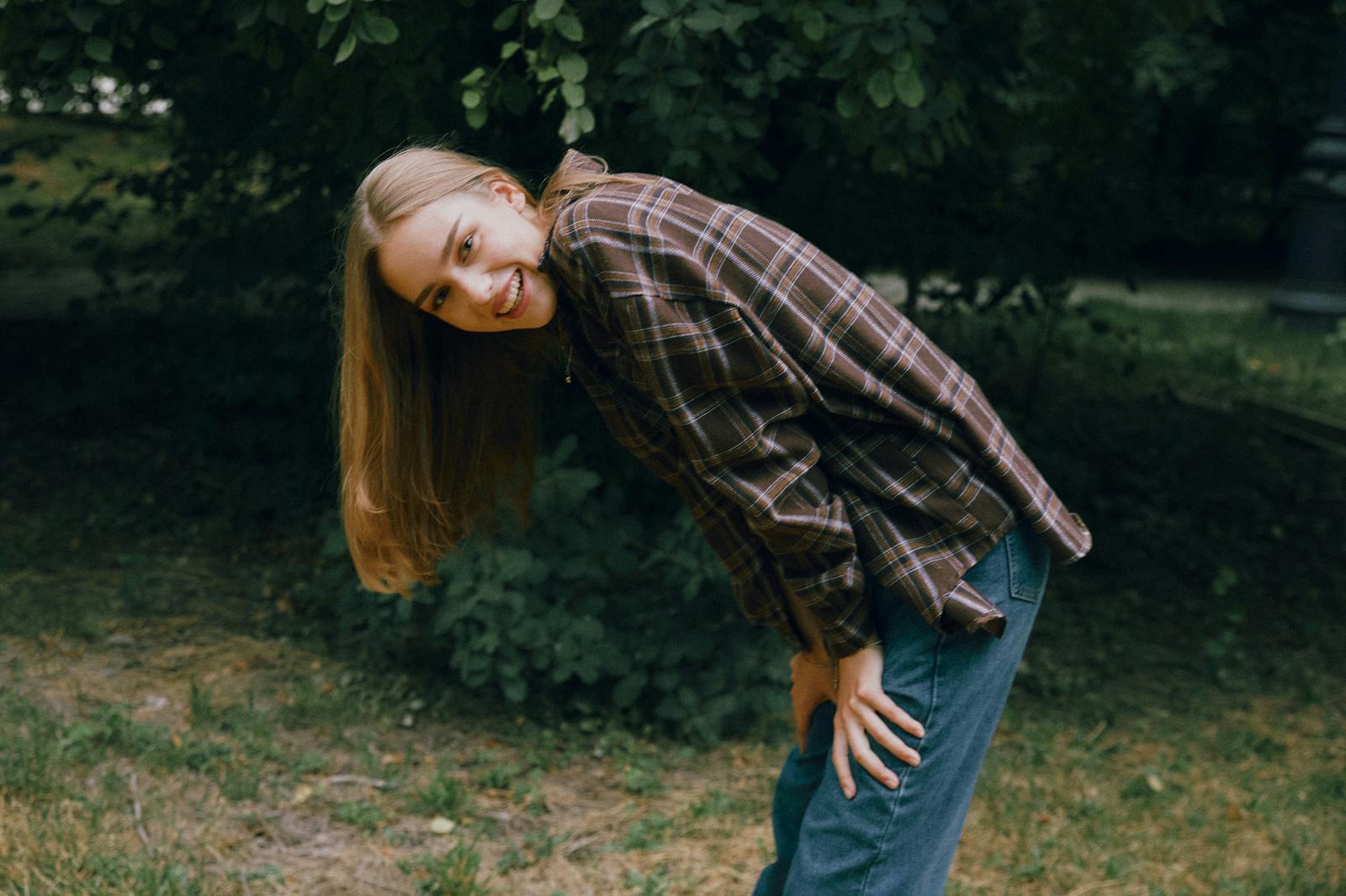 A joyful young woman with long hair bends and smiles outdoors in casual attire.