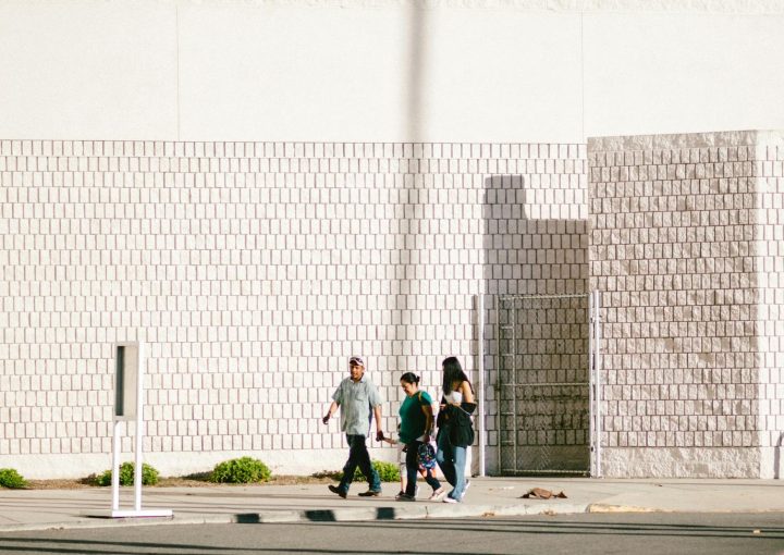 A group walks past a textured brick wall in bright daylight, casting long shadows.