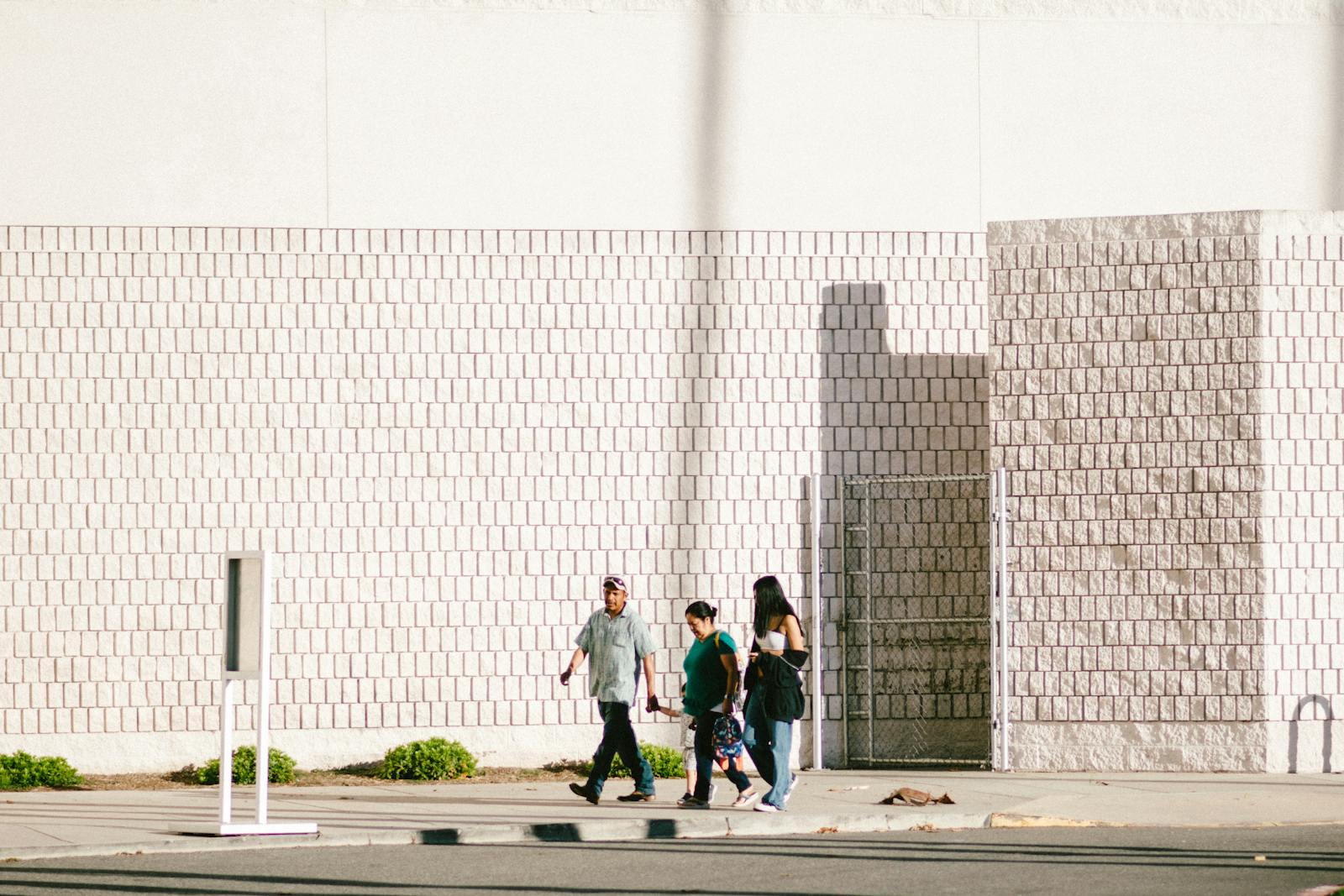 A group walks past a textured brick wall in bright daylight, casting long shadows.