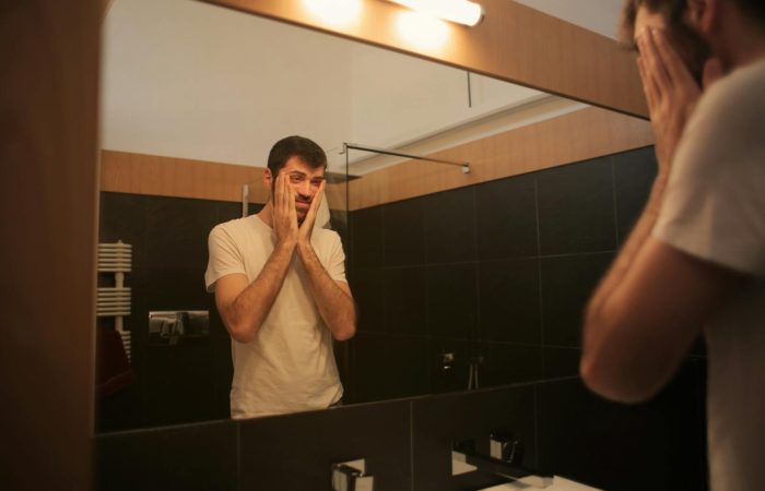 Man in white t-shirt reflects in bathroom mirror, appearing thoughtful and concerned.
