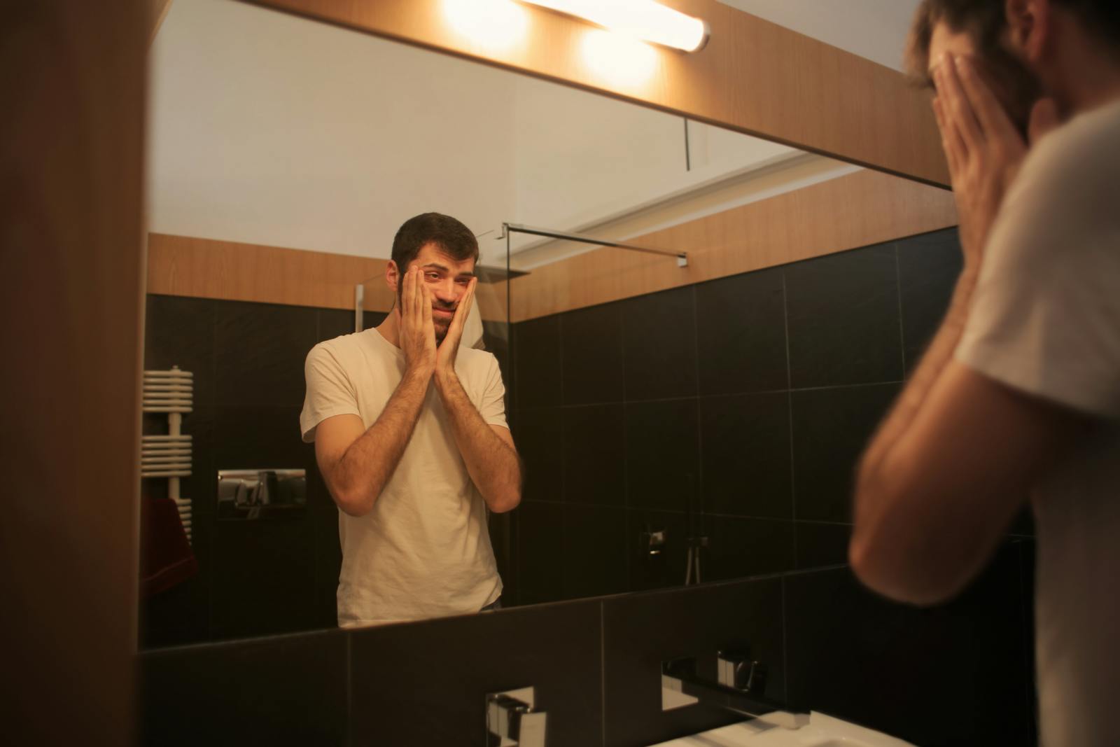 Man in white t-shirt reflects in bathroom mirror, appearing thoughtful and concerned.