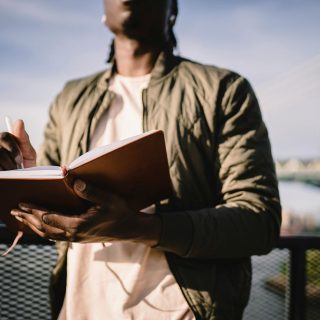 From below of crop anonymous African American male in casual clothes standing near terrace fence with open journal and pen behind bridge over river in daylight