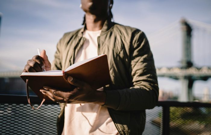 From below of crop anonymous African American male in casual clothes standing near terrace fence with open journal and pen behind bridge over river in daylight