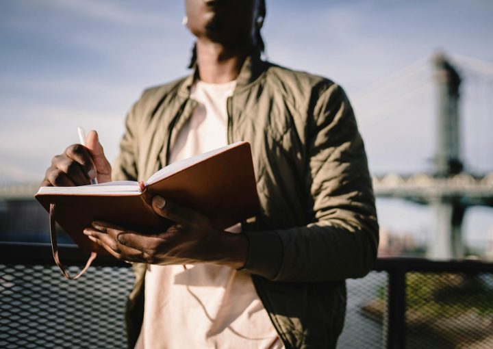 From below of crop anonymous African American male in casual clothes standing near terrace fence with open journal and pen behind bridge over river in daylight