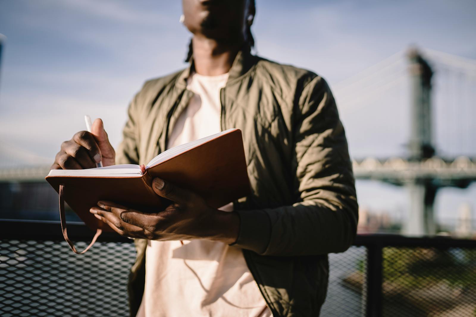 From below of crop anonymous African American male in casual clothes standing near terrace fence with open journal and pen behind bridge over river in daylight