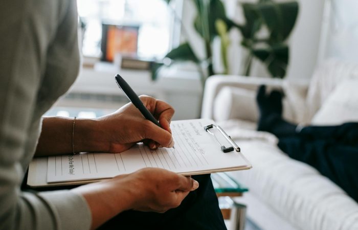 Unrecognizable ethnic female therapist taking notes on clipboard while filling out form during psychological appointment with anonymous client lying on blurred background