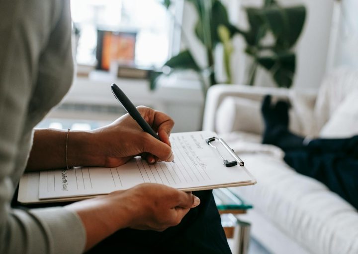 Unrecognizable ethnic female therapist taking notes on clipboard while filling out form during psychological appointment with anonymous client lying on blurred background
