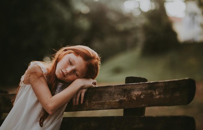 A young girl sleeps peacefully on a wooden bench outdoors, surrounded by nature.