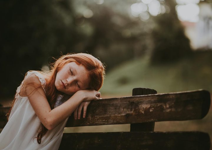 A young girl sleeps peacefully on a wooden bench outdoors, surrounded by nature.