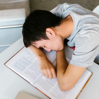 A young man sleeping on a white table with an open textbook, depicting study fatigue.