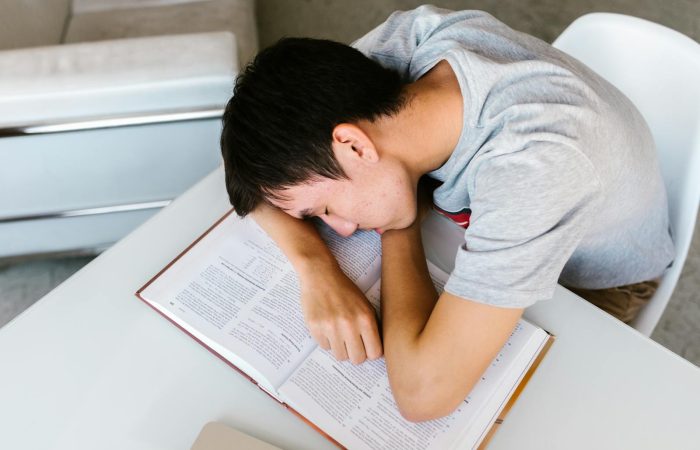 A young man sleeping on a white table with an open textbook, depicting study fatigue.