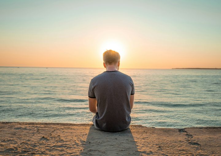 man sitting on sand front of sea during golden hour